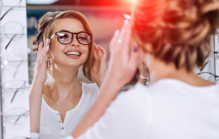 A young woman smiling as she tries on new prescription glasses at the optometrist office.