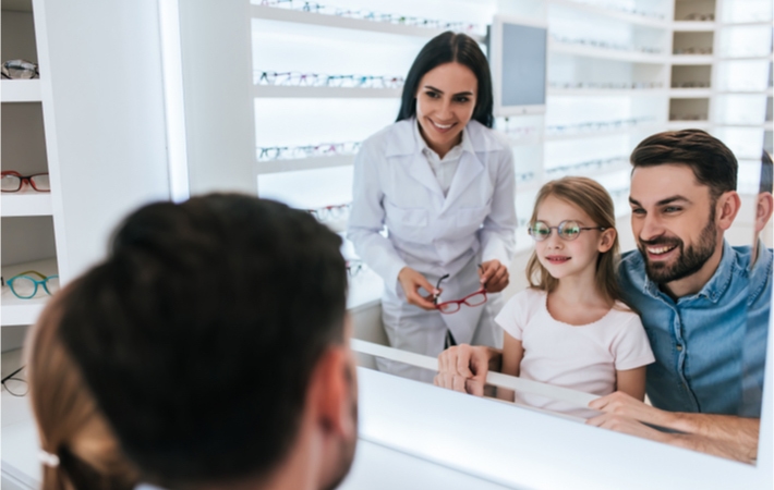 A smiling dad and his daughter being assisted by an optician as she tries on frames.