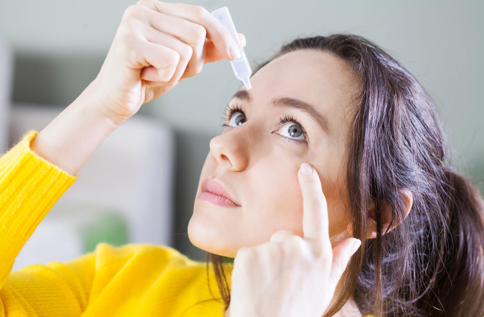 A woman holding a small bottle of eye drops in her right hand and putting them on her left eye.
