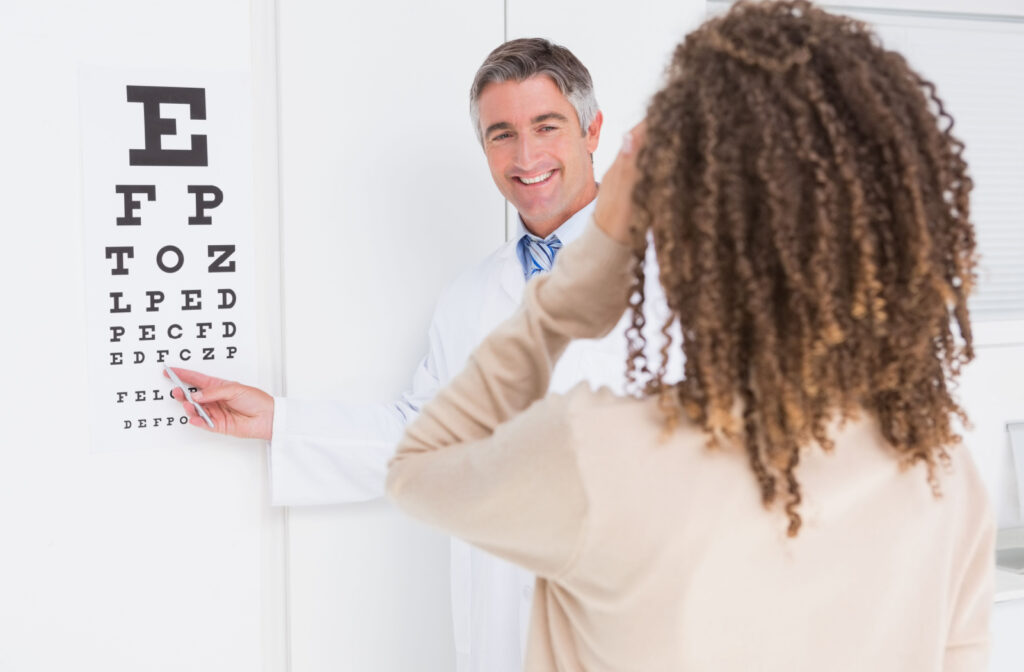 A woman standing and gazing forward while her eye doctor gestures towards an eye chart during an eye exam.