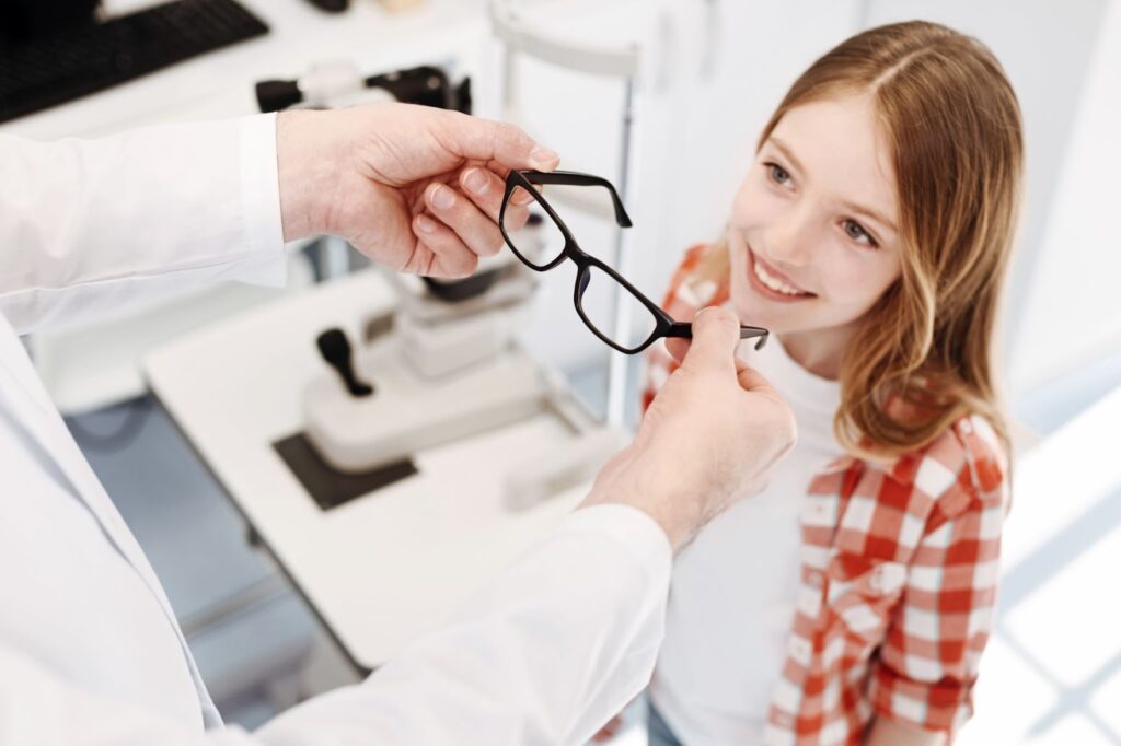 A young girl smiling while an optician holds a pair of glasses in front of her to try.