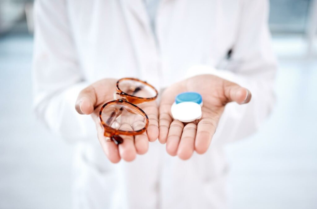 An optician in a white coat with outstretched palms holding eyeglasses in the right palm and a contact lens case in the left palm.