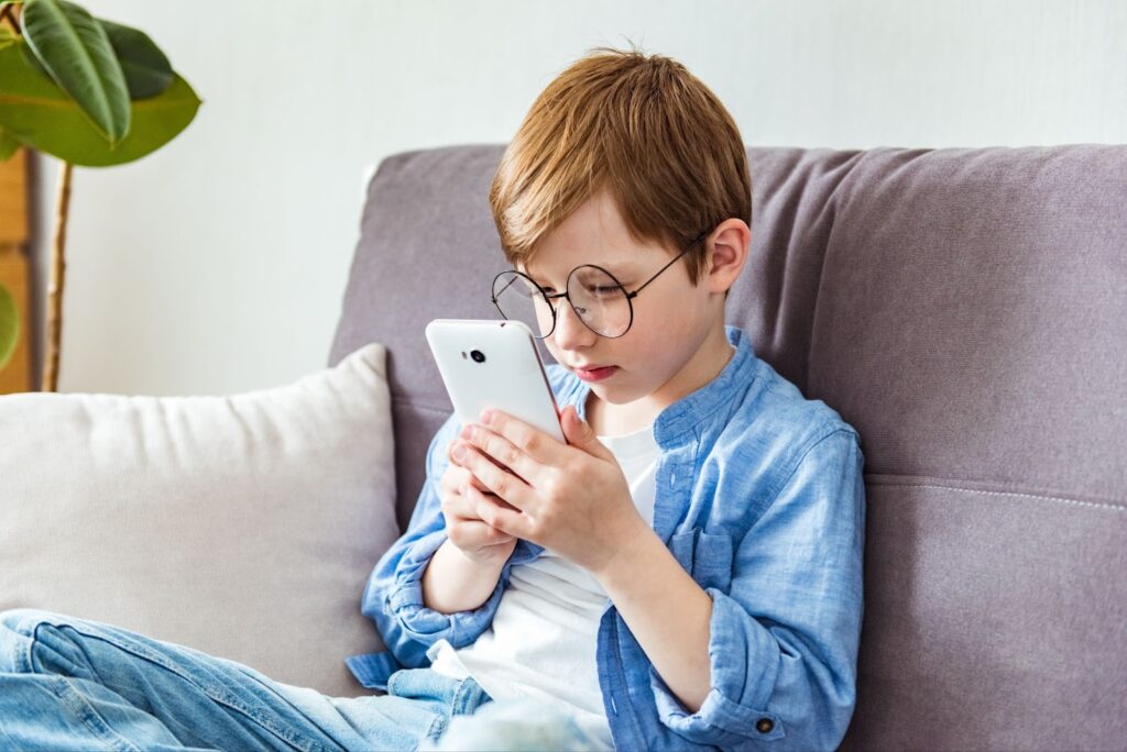 A young child wearing glasses sits on a couch and watches something on their tablet.