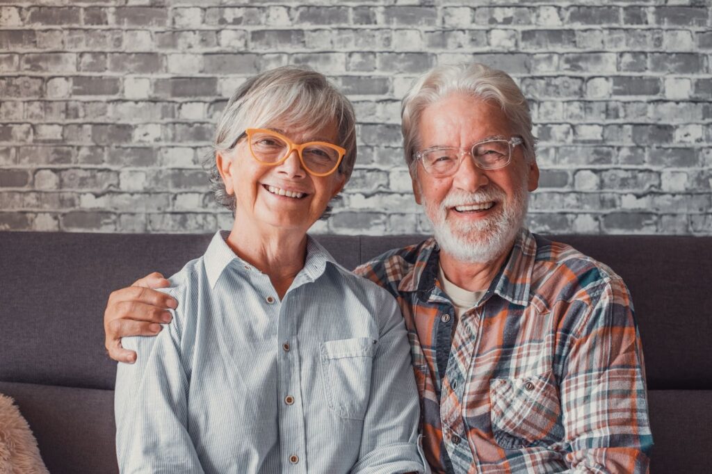 An older couple wearing acetate glasses sitting together smiling on the couch.
