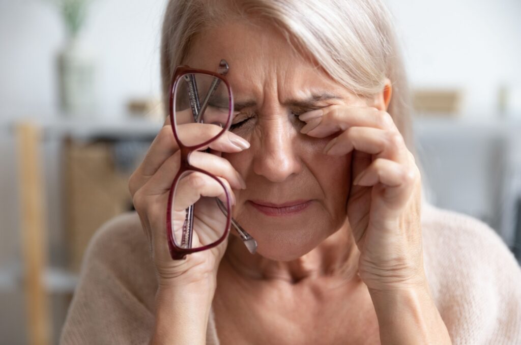 An older adult rubbing their eyes from dry eye syndrome while holding a pair of glasses in their hands.