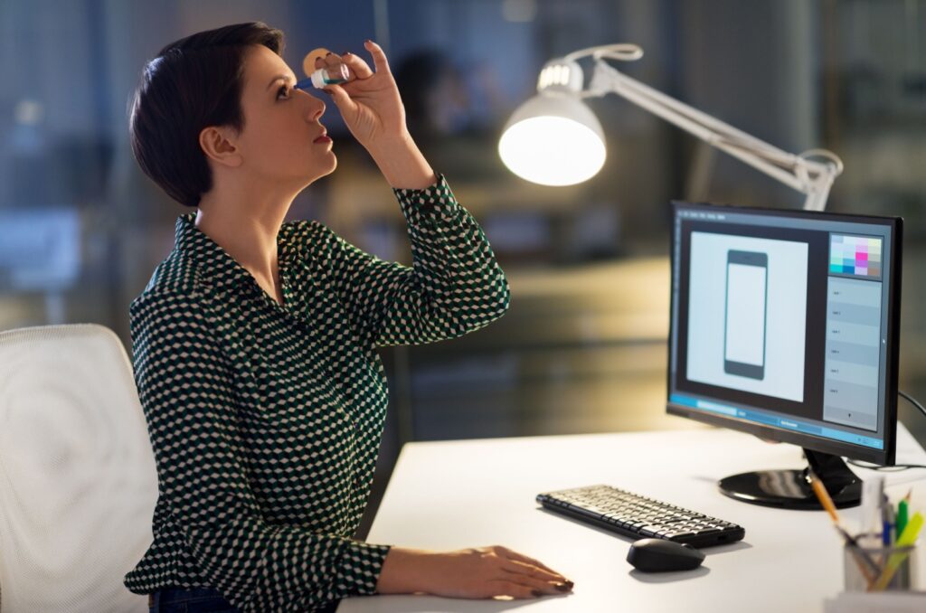 An office worker sitting at a desk with a laptop and lamp and putting eye drops in their eyes.