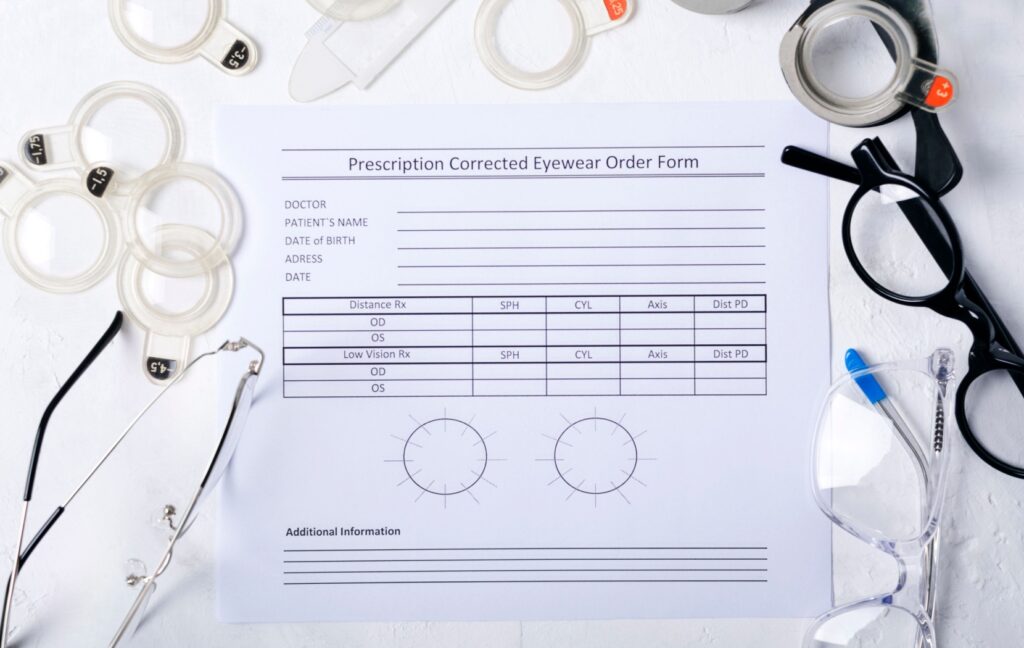 A blank eyeglass prescription form sits on a table as several pairs of eyeglasses and vision testing equipment are arranged around it.
