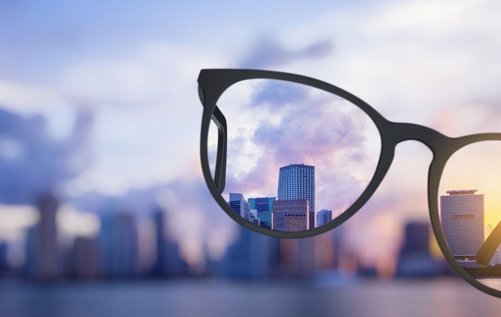 A blurry city skyline is brought into focus with a pair of corrective eyeglasses held up in front of the camera.