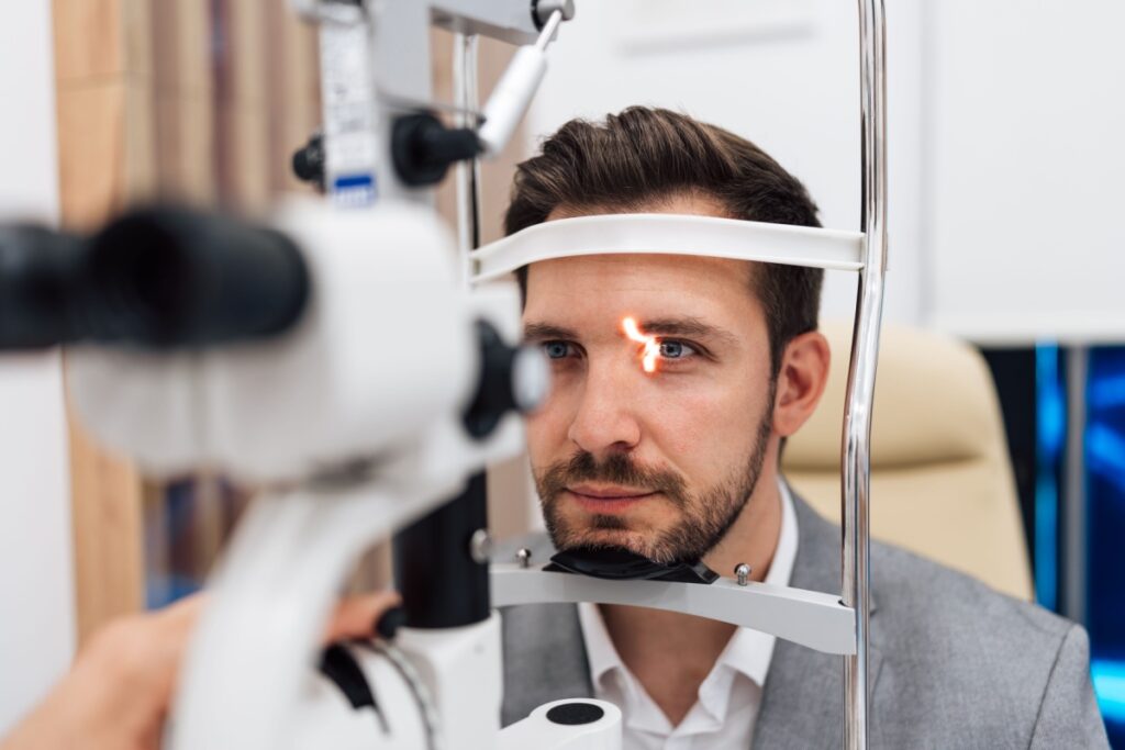 An adult man sitting at a slit lamp while an eye doctor examines his eye.