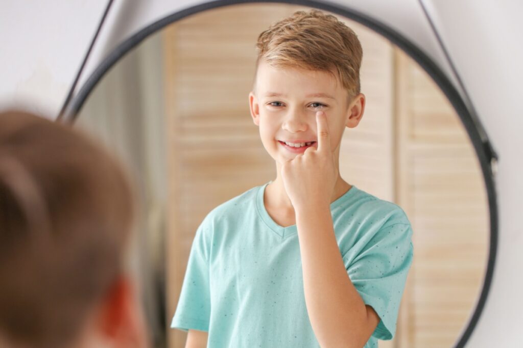 A young boy standing in front of a mirror and pointing to his eye with a smile.