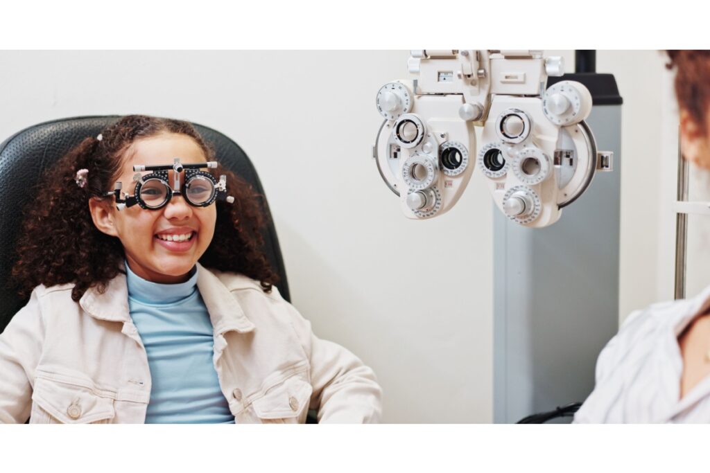 A young girl smiling during a vision test while wearing trial frames in an optometry exam room.