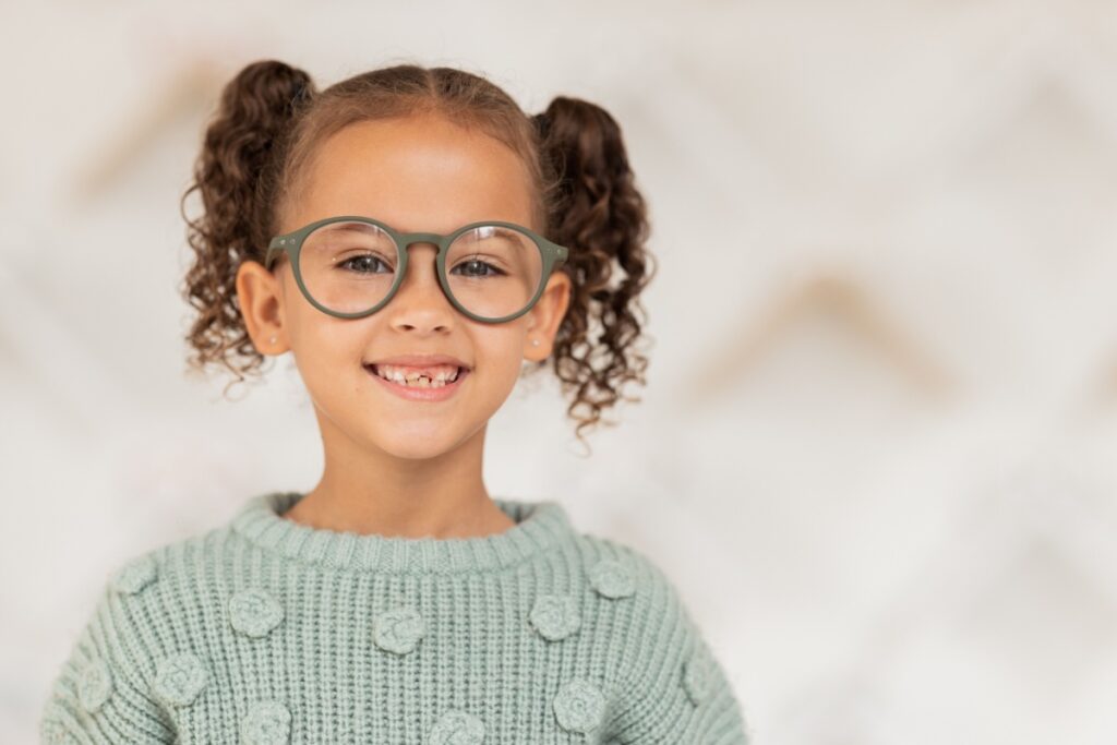 A young girl with curly hair wearing round green glasses and a knitted sweater, smiling at the camera.